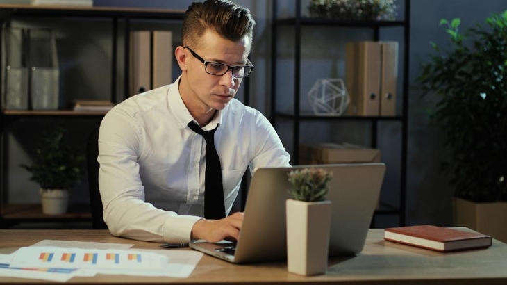 A man in glasses and a shirt focused on his laptop, engrossed in his work.