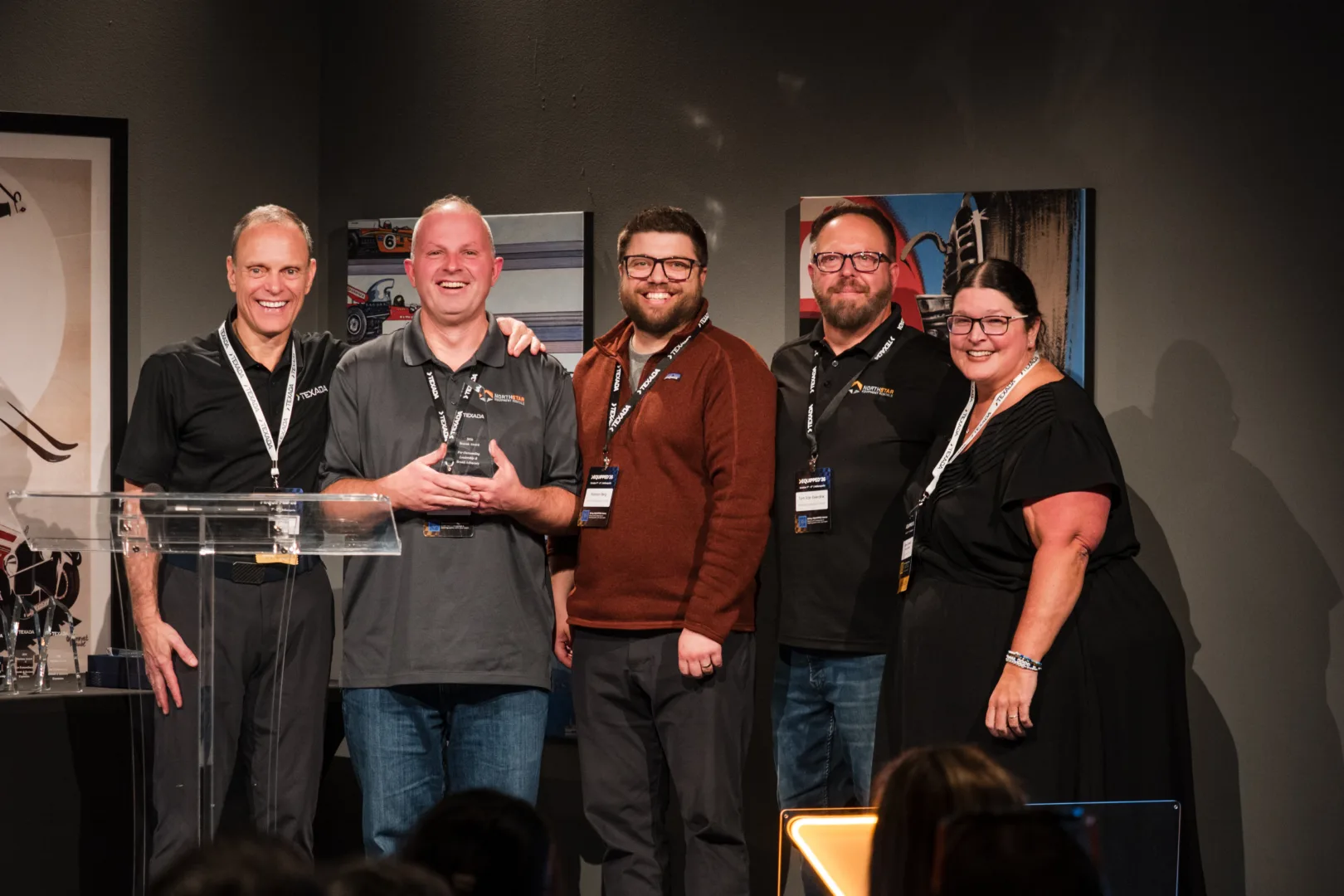 Five people stand on a stage posing for a photo, one person holding a glass award, all wearing event lanyards.